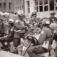 Iconic image of working German Shepard dog sitting on a driving cart surrounded by firefighters during 911 response.