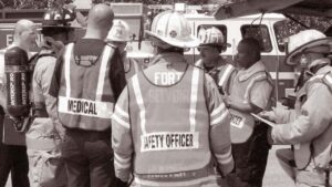 Group of firefighters and other responders from different agencies standing together talking. Some are wearing vests identifying them as medical, safety officer.